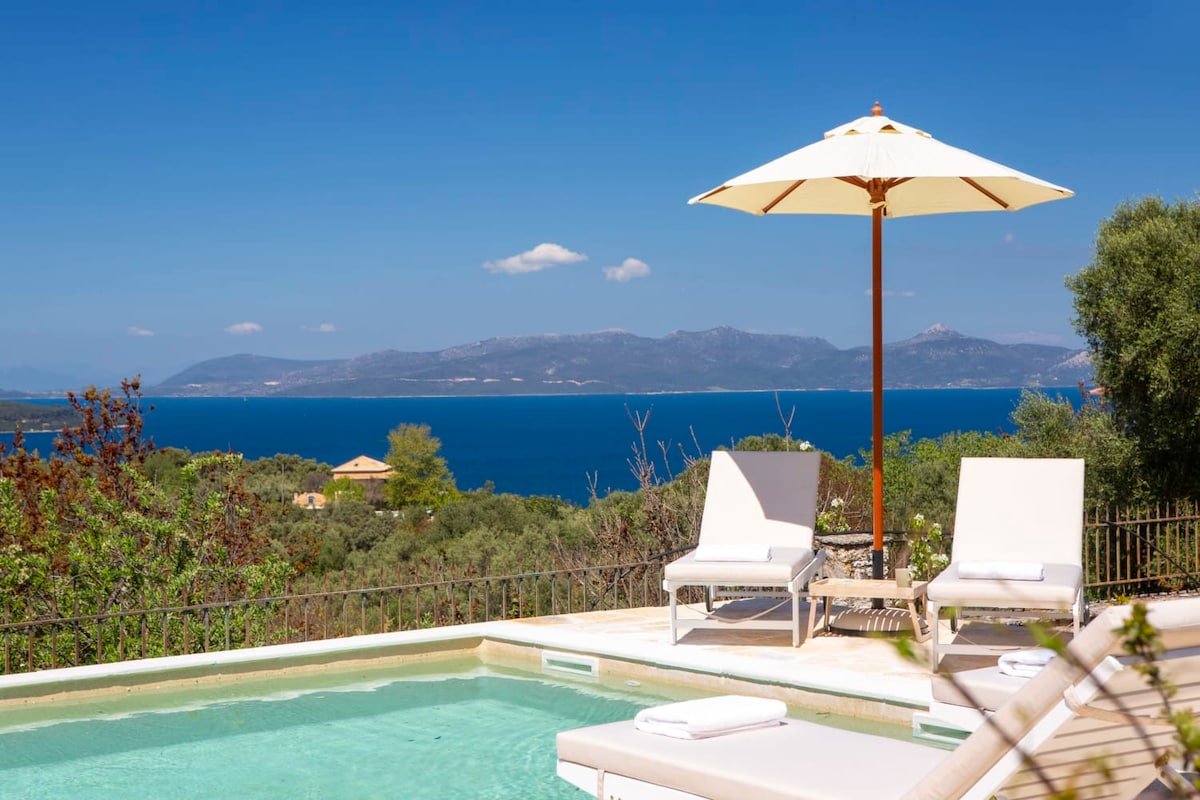 A private pool area is visible, featuring two sun loungers positioned under a large umbrella. The crystal-clear water reflects the vibrant blue of the sea in the background, with lush greenery surrounding the space, creating a serene and tranquil atmosphere.