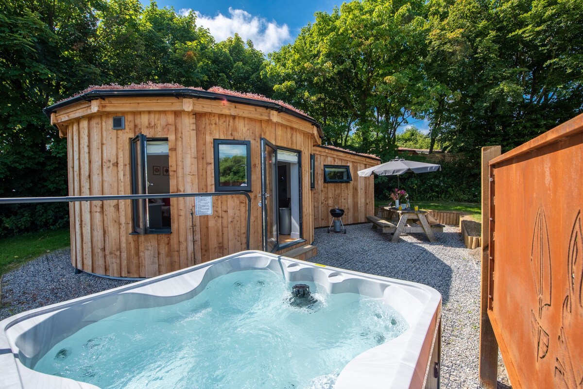 A round wooden structure is surrounded by lush greenery, featuring large windows and a private outdoor area. A hot tub is positioned nearby, with a picnic bench and a charcoal BBQ setup partially visible on the gravel surface.