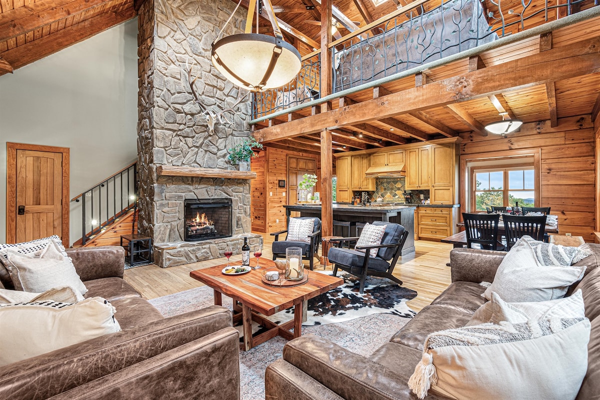 The open living space features a large stone fireplace as a focal point, surrounded by comfortable leather sofas. A wooden coffee table sits at the center, with decorative items placed atop. A view of the kitchen and wooden beams in the ceiling adds warmth to the setting.