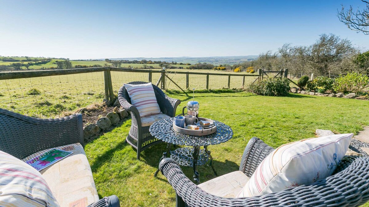 A small garden area with woven seating accommodates outdoor relaxation. A round table sits at the center, adorned with a tray and refreshments. The fenced space offers a view of rolling hills and open fields, creating a peaceful backdrop for guests to enjoy.