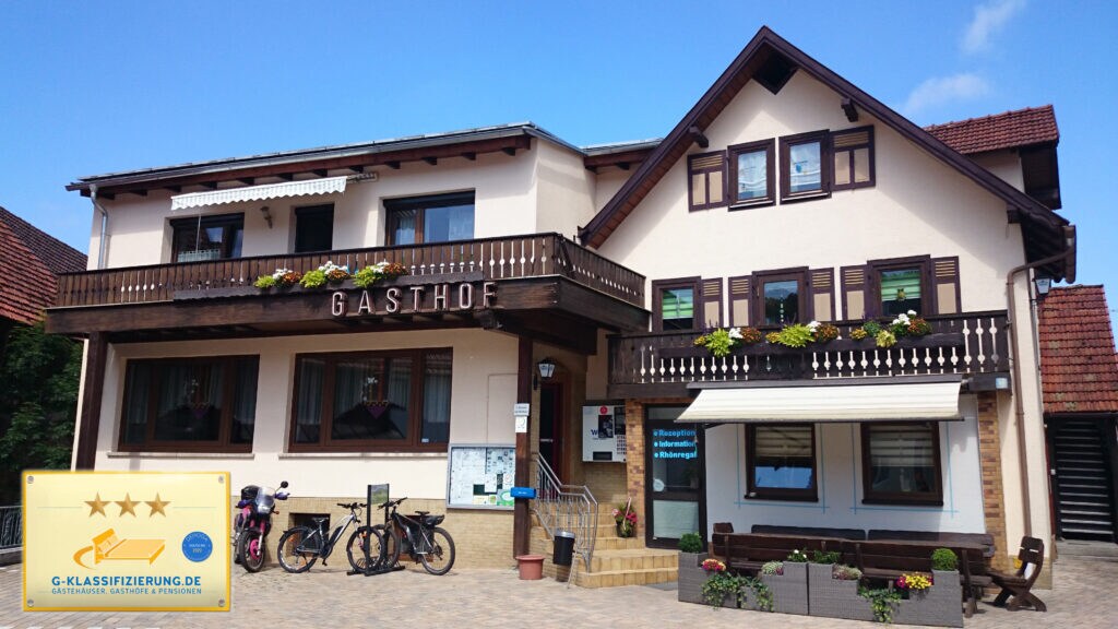 A three-star guesthouse is displayed with a welcoming facade, featuring a wooden balcony adorned with flower boxes. Bicycles are positioned nearby, and outdoor seating is visible at the front. The building's architecture combines traditional elements with modern details, set against a clear blue sky.