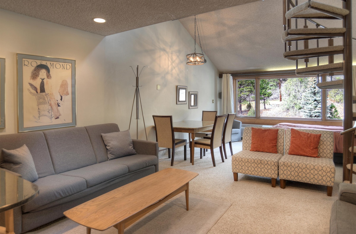 A comfortable living space features a gray sofa facing a light wooden coffee table. A dining area includes a square table with four chairs. Natural light fills the room through large windows, revealing views of the outdoors. A spiral staircase leads to the lofted area.