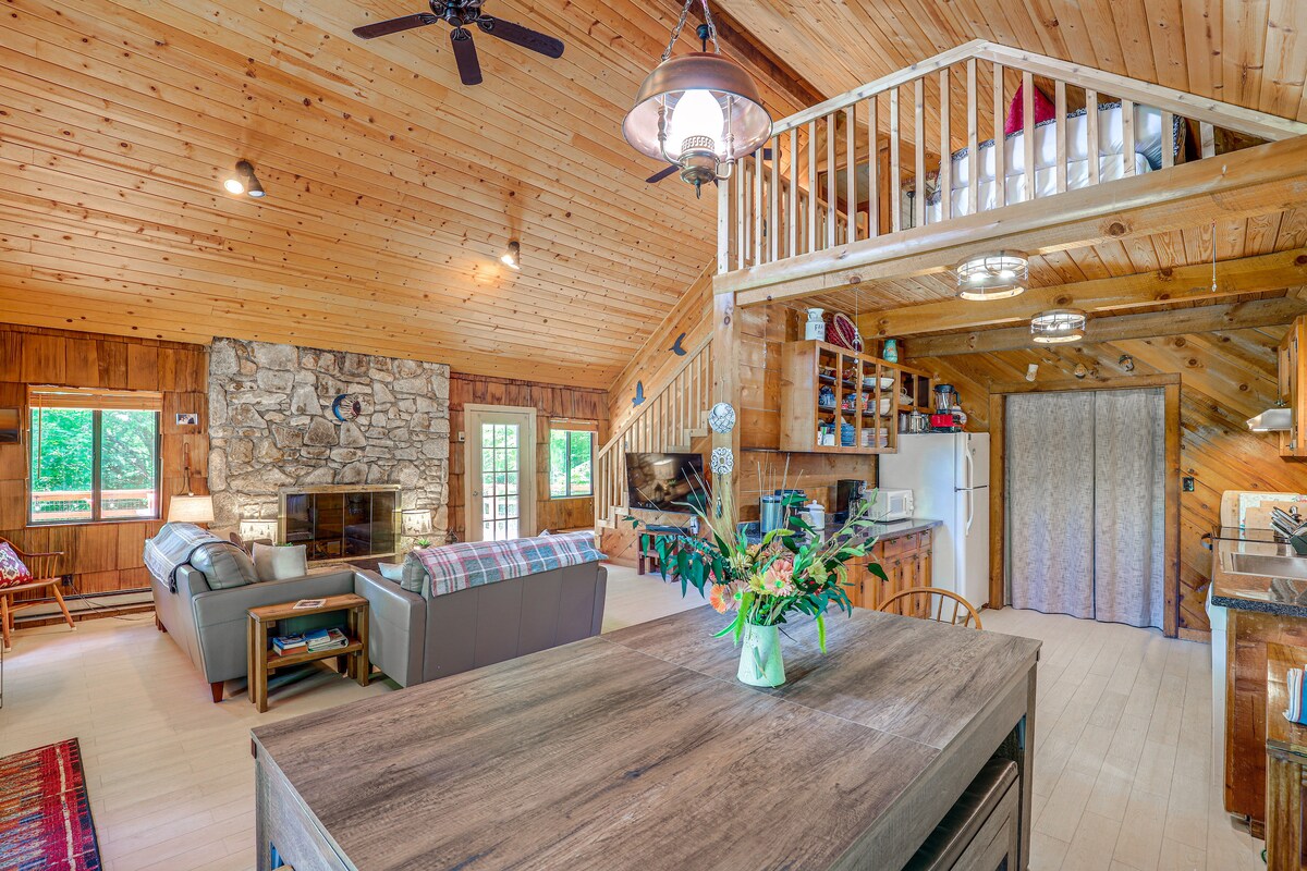 A spacious living area features wood-paneled walls and a vaulted ceiling. Comfortable seating arrangements surround a stone fireplace, with large windows allowing natural light to fill the space. A wooden dining table is positioned centrally, complemented by the view of an adjacent kitchen area.