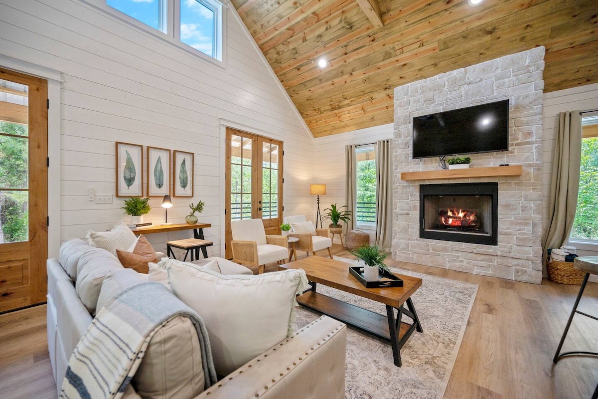 A bright living area is showcased, featuring a stone fireplace that adds warmth. Light wood beams line the ceiling, while large windows invite natural light. The space includes a comfortable couch and two armchairs around a central coffee table, creating a welcoming gathering spot.