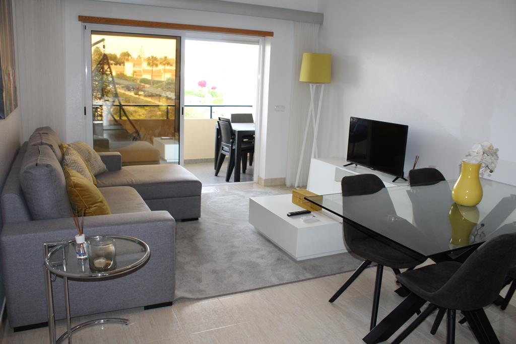 A modern living area features a gray sectional sofa with yellow accent pillows. A glass dining table is surrounded by dark chairs. Natural light filters in from the sliding glass doors that open to a small balcony, enhancing the spacious appearance of the room.
