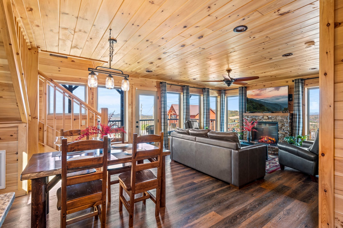 An open living area features large windows that provide expansive mountain views. Comfortable leather seating surrounds a stone fireplace, and a wooden dining table accommodates multiple guests. Warm wood paneling and a polished floor enhance the rustic charm of the space.