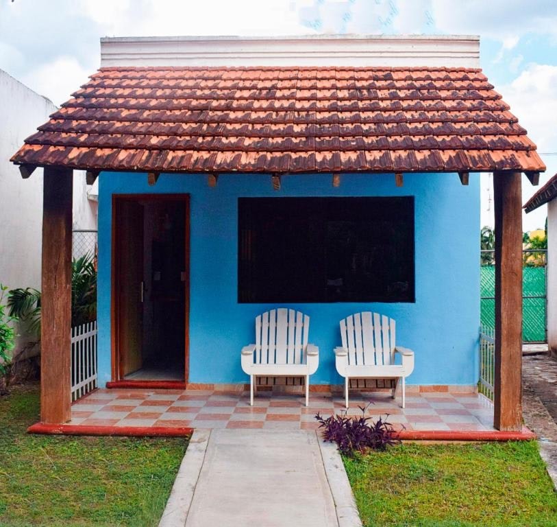 A charming blue villa exterior features a traditional gabled roof with terracotta tiles. Two white Adirondack chairs are positioned on a tiled porch, inviting relaxation. The pathway leads to a door framed by greenery, enhancing the welcoming atmosphere of the property.