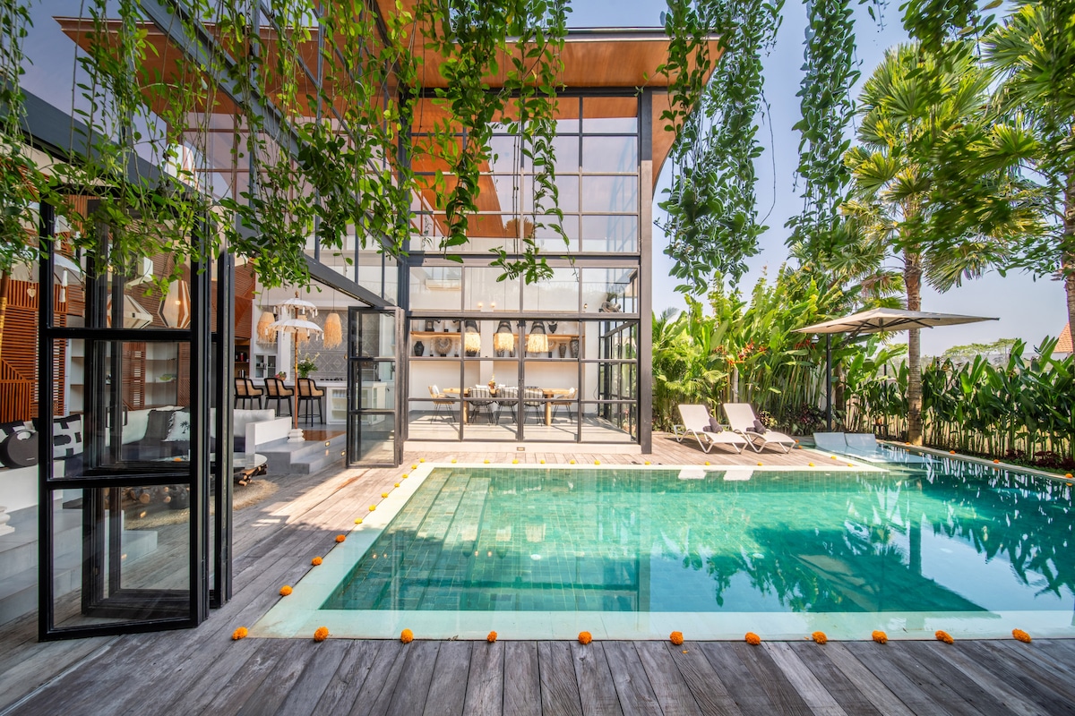 The image showcases a modern villa with a crystal-clear swimming pool surrounded by wooden decking. Lush greenery and tropical plants frame the pool area, while large glass doors provide a view into the spacious living area behind. Comfortable loungers are positioned near the water.