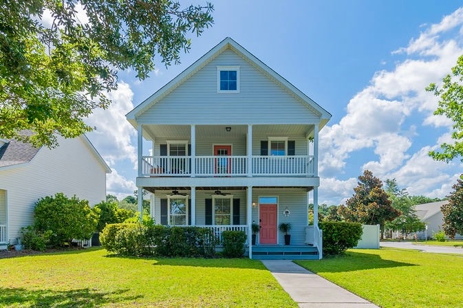 A charming two-story house is presented with a light exterior and a welcoming orange door. The covered porch features white railings and multiple seating areas. Lush greenery surrounds the home, enhancing its outdoor space, while a clear blue sky with scattered clouds creates a pleasant backdrop.