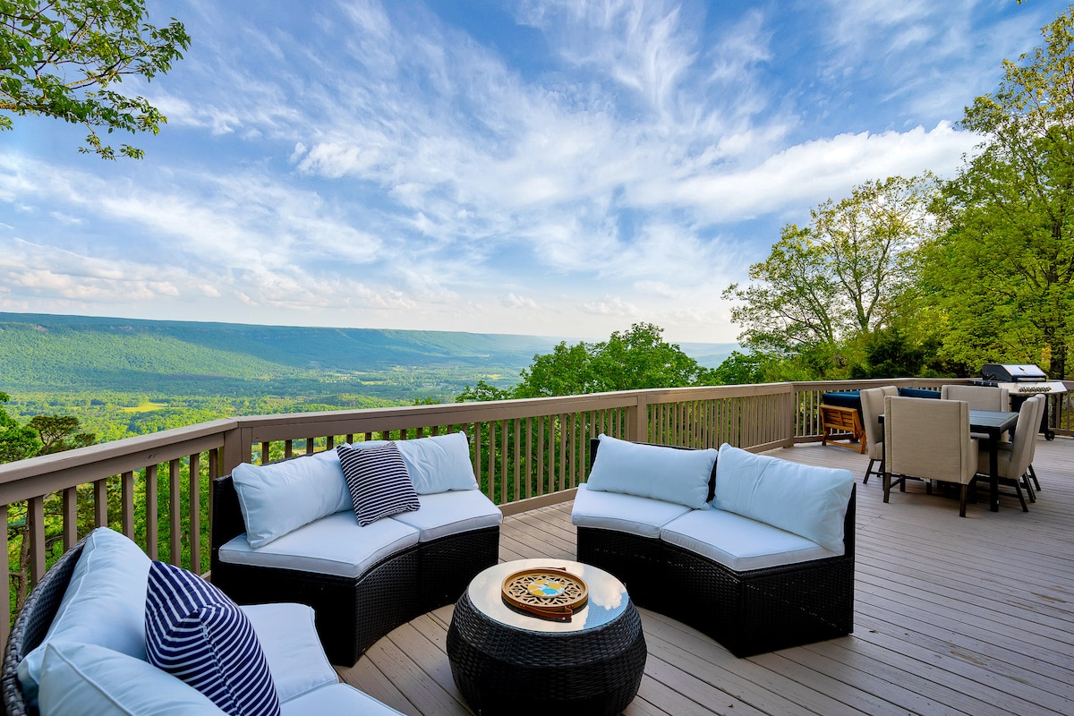 A spacious outdoor deck features comfortable seating arrangements with plush cushions, offering breathtaking views of the surrounding landscape. A circular table sits at the center, surrounded by lush greenery and expansive skies, presenting a perfect setting for relaxation and enjoying nature.