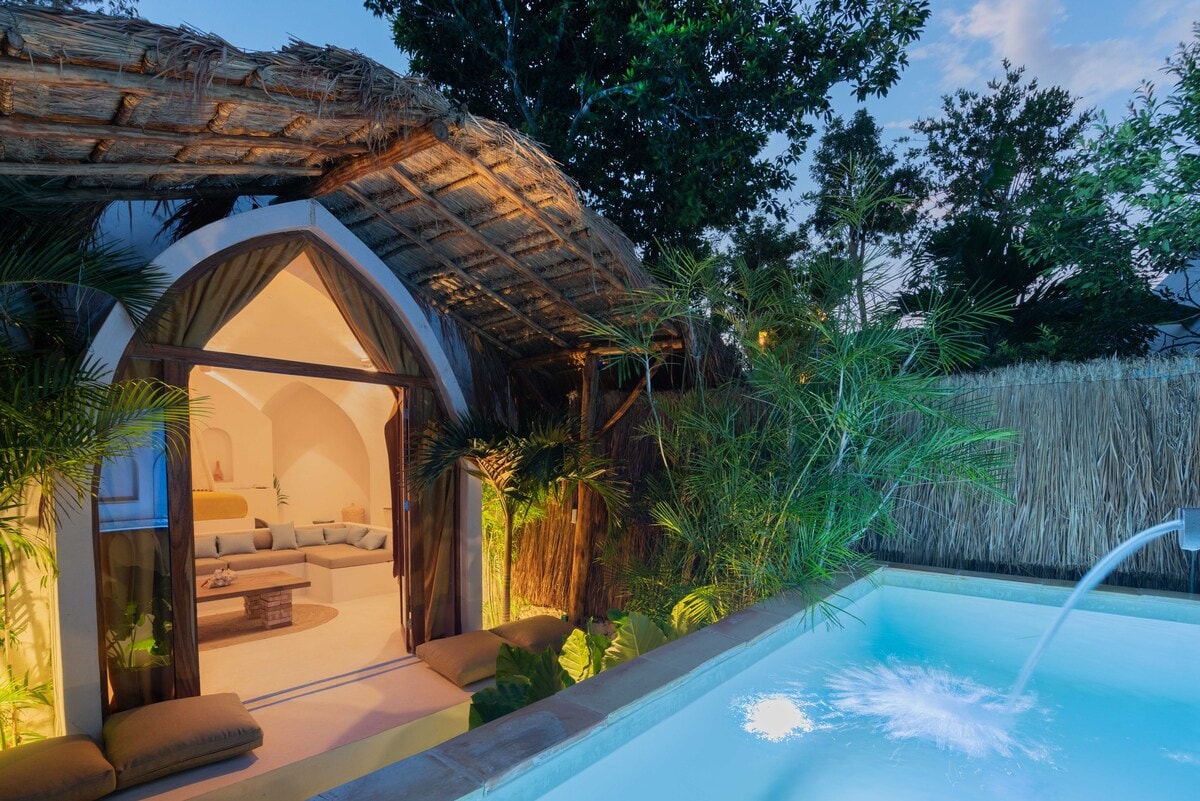 An outdoor area features a small pool, with water gently cascading from a spout. Lush greenery surrounds the space, while a thatched roof offers partial cover. Soft lighting emanates from the interior, visible through large glass doors that open into a cozy living space.