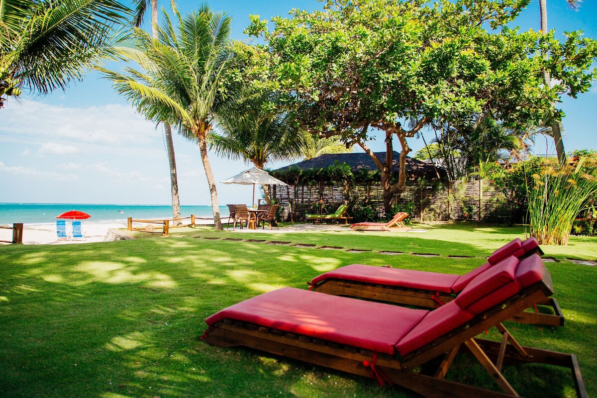 Sun loungers with red cushions are positioned on a lush green lawn, facing the beach. Palm trees and tropical foliage provide shade, while a glimpse of the ocean is visible in the background. A wooden structure is partially seen behind the trees, contributing to the tranquil outdoor setting.