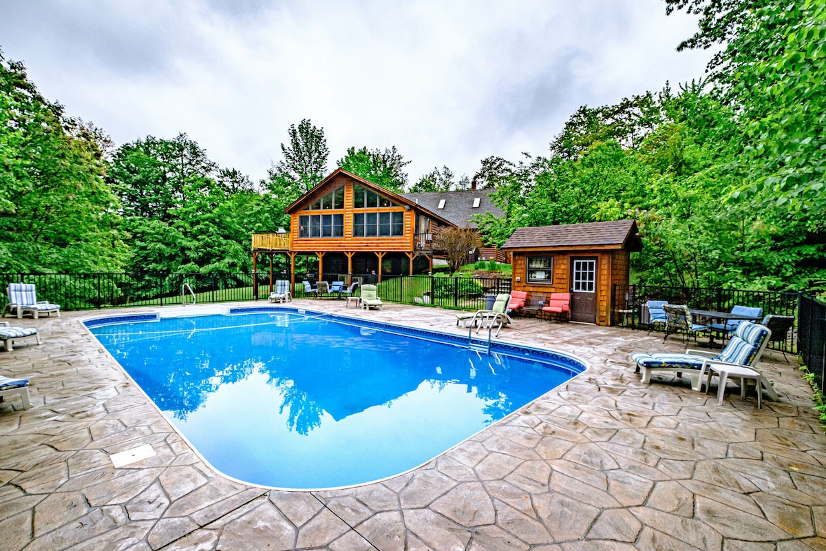 A spacious in-ground pool is framed by a stone patio, surrounded by lush greenery. The pool area includes lounge chairs and an outdoor structure, providing shade. A large wooden building with multiple windows overlooks the pool, enhancing the inviting atmosphere for outdoor gatherings.