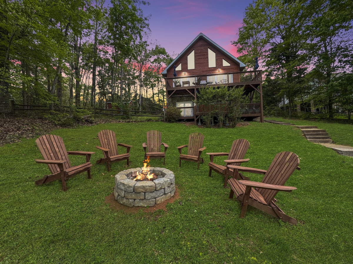 An inviting outdoor fire pit is surrounded by eight wooden Adirondack chairs on a landscaped lawn. In the background, the cottage stands among trees, featuring a spacious deck that overlooks the serene setting. Soft dusk light enhances the tranquil atmosphere.