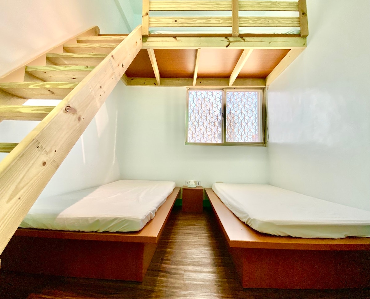 A simple sleeping area is observed, featuring two elevated wooden platforms with mattresses. A ladder leads to a lofted space above. A window with a latticed design allows natural light to enter, illuminating the minimalist decor of the room.