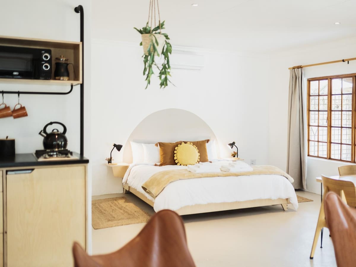 A cozy room features a king-size bed with a decorative round pillow and light bedding against a white wall. A kitchenette with a microwave is visible, along with wooden dining chairs and a woven rug, creating a comfortable, functional space.