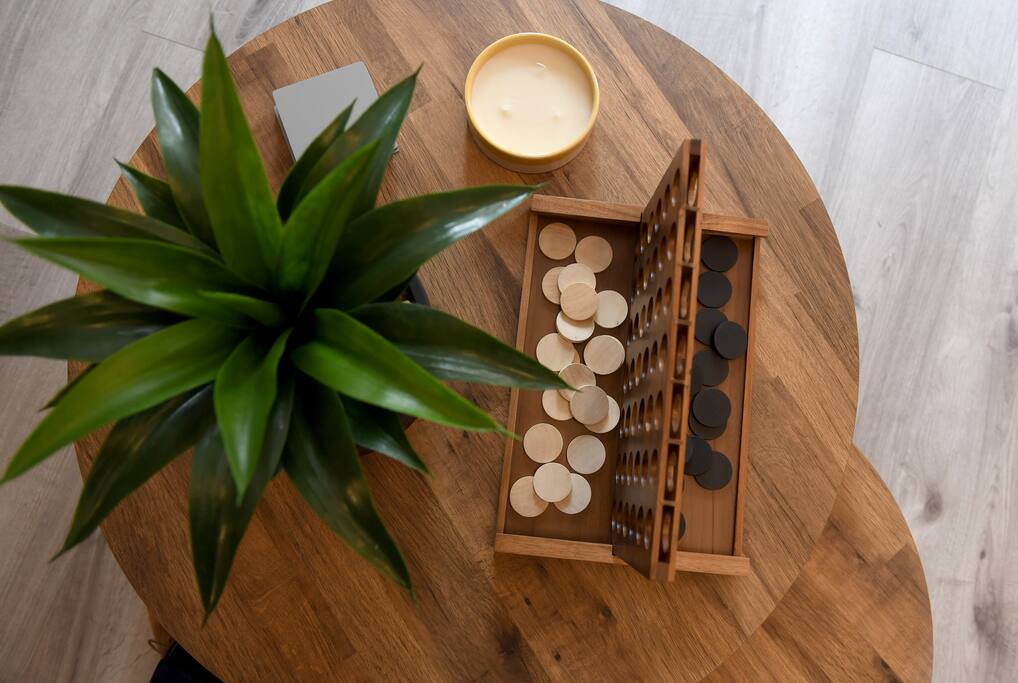 A round wooden coffee table features a thoughtful arrangement of a game set with black and white pieces, alongside a small candle and a bowl. A green plant adds a touch of life, complementing the cozy atmosphere.