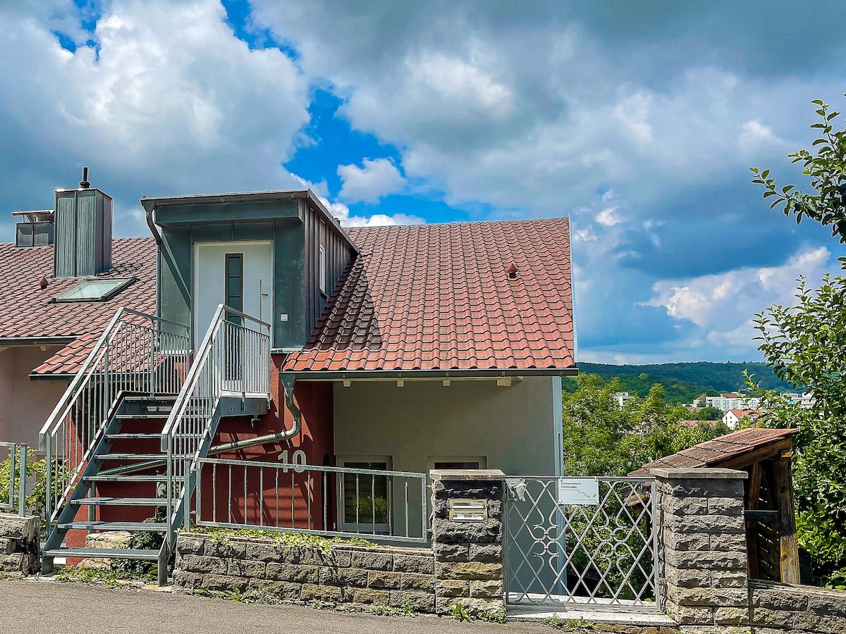 The exterior features a two-story building with a sloped roof and red tiles. A staircase leads to the main entrance, framed by a stone wall and railing. Lush greenery and a blue sky filled with clouds add to the overall setting.
