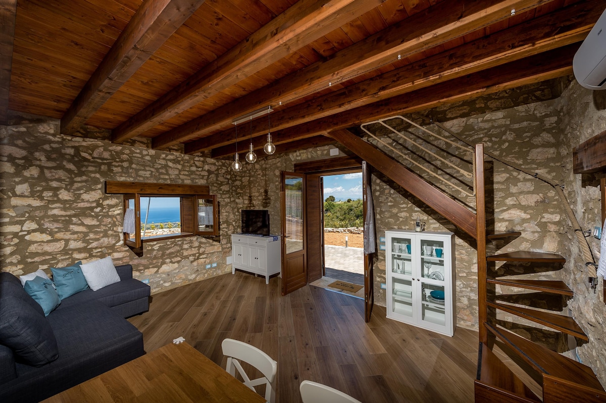 An open living area features rustic stone walls and wooden beams. A comfortable dark sofa is positioned beside a light wooden dining table. A staircase leads to the upper level, while a door opens to a view of the countryside. A television is mounted on the wall.