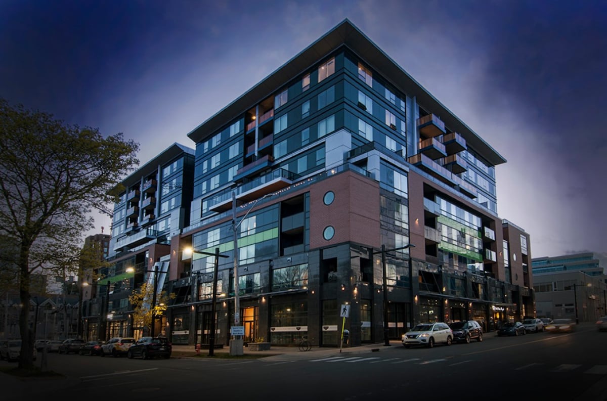 The contemporary multi-story building features a sleek design with large windows and balconies. Streetlights illuminate the facade, enhancing its modern architecture. The surrounding area includes parked cars and tree-lined sidewalks, contributing to a vibrant urban environment.