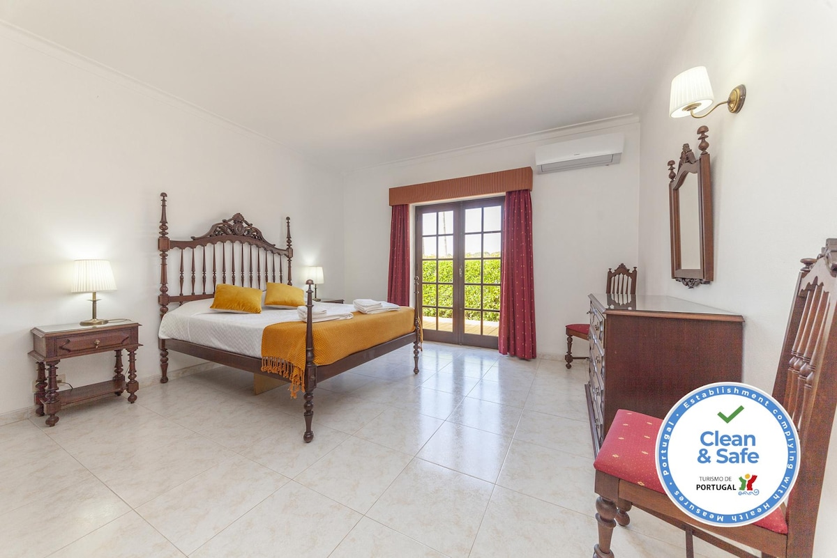 The spacious bedroom features a double bed with an orange throw. A wooden dresser and matching chair are positioned near the window, which is framed by red curtains. Soft light from the lamps enhances the bright, airy atmosphere.