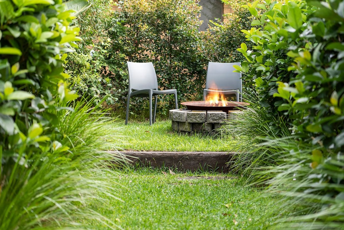 A serene outdoor space is framed by lush greenery, featuring two gray chairs positioned around a circular fire pit. The grass is well-maintained, creating a welcoming area for relaxation and gatherings. The fire pit is surrounded by stone, enhancing the cozy atmosphere.