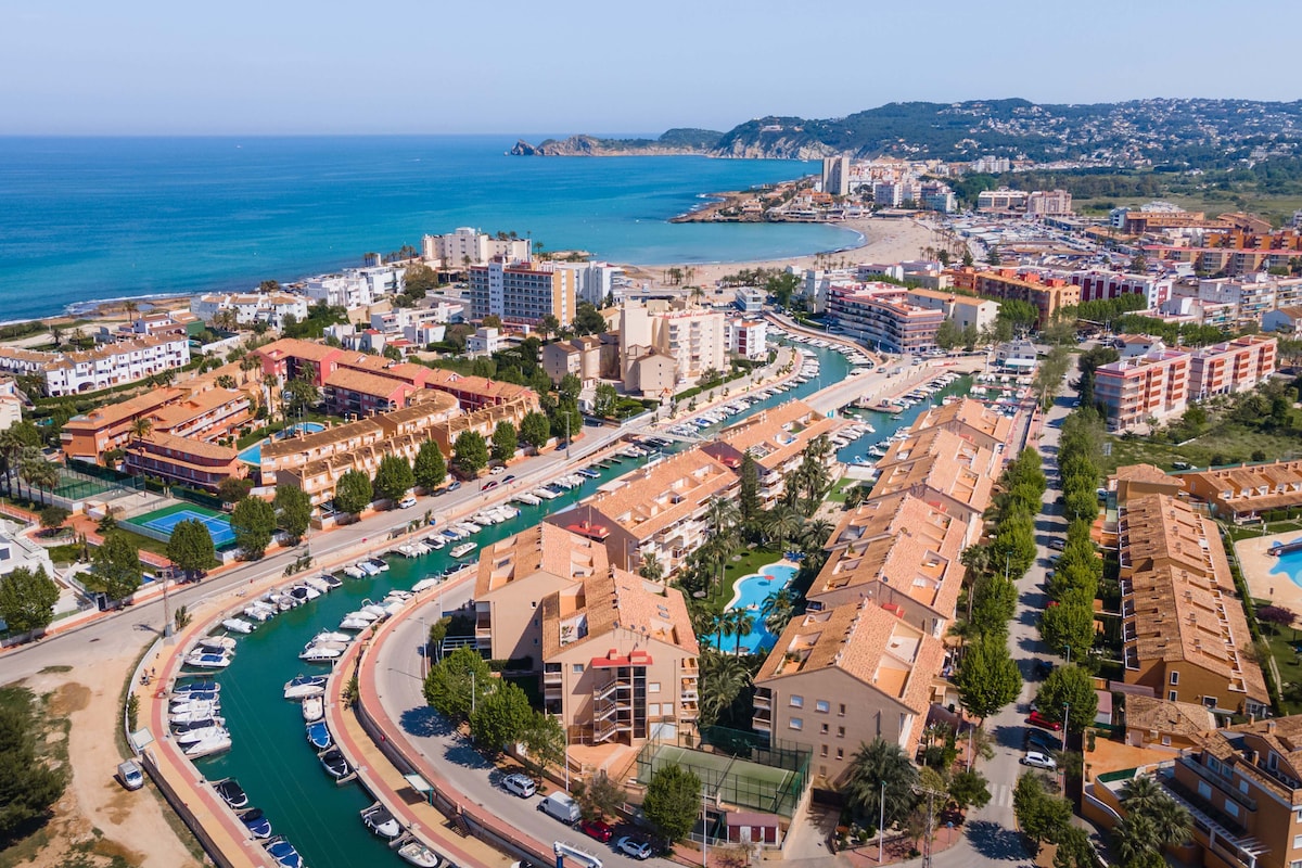 Aerial view of the scenic coastline featuring sandy beaches and a marina filled with boats. The image captures a mix of colorful buildings and green spaces, highlighting the proximity of the apartment complex to both the coast and well-maintained gardens.