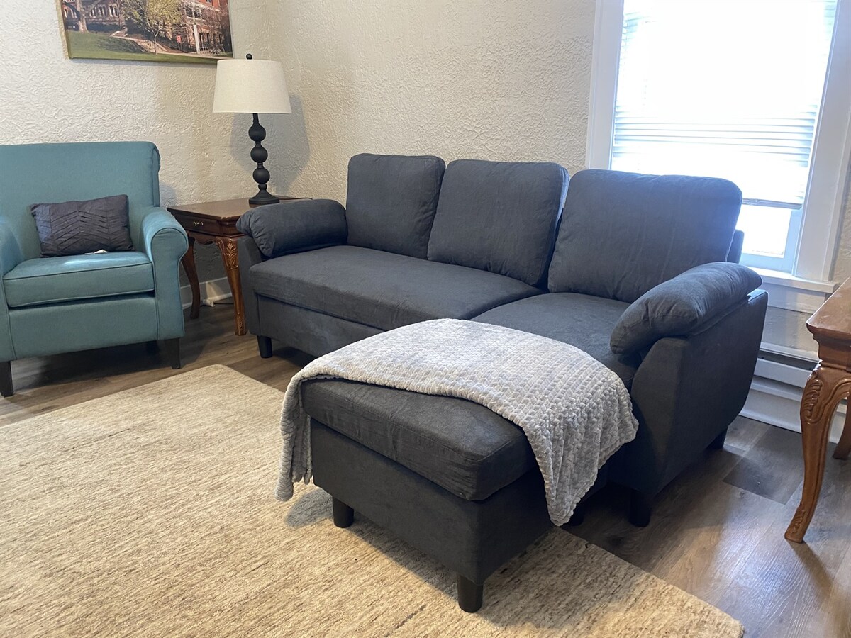 The living area features a comfortable sectional sofa and a lounge chair, inviting relaxation. A light-colored throw blanket rests on the sofa. A wooden side table and a floor lamp offer additional lighting, while natural light filters through the nearby window.