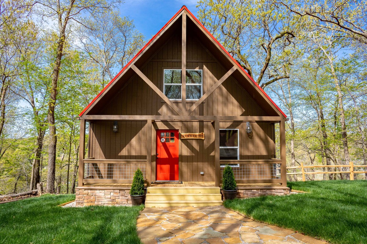 The exterior of a rustic cabin is showcased, featuring a wood-paneled facade and a bright red front door. Natural stone steps lead up to the entrance, surrounded by a well-manicured green lawn and mature trees. Large windows are visible, enhancing the cabin's charm.