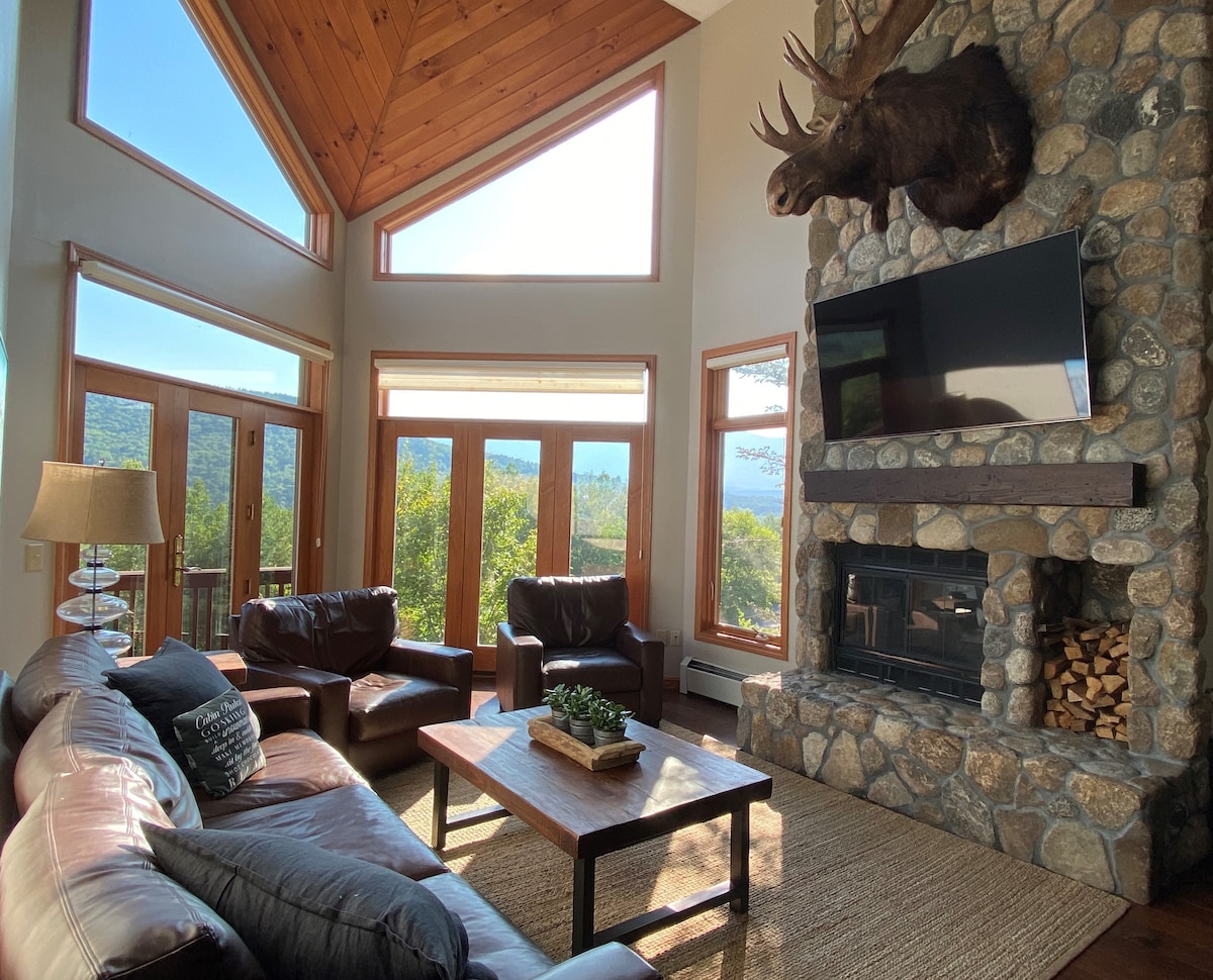 An expansive living area features a stone fireplace and large windows that provide ample natural light and views of the surrounding landscape. Leather seating surrounds a wooden coffee table, while a decorative plant adds a touch of greenery. The rustic wooden ceiling enhances the room's inviting ambiance.