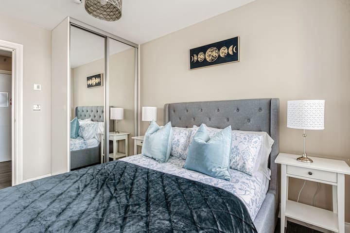 Elegante dormitorio con cabecera gris acolchada, armario con espejos, obras de arte modernas en fase lunar y lujosa ropa de cama en tonos azul marino y azul claro.