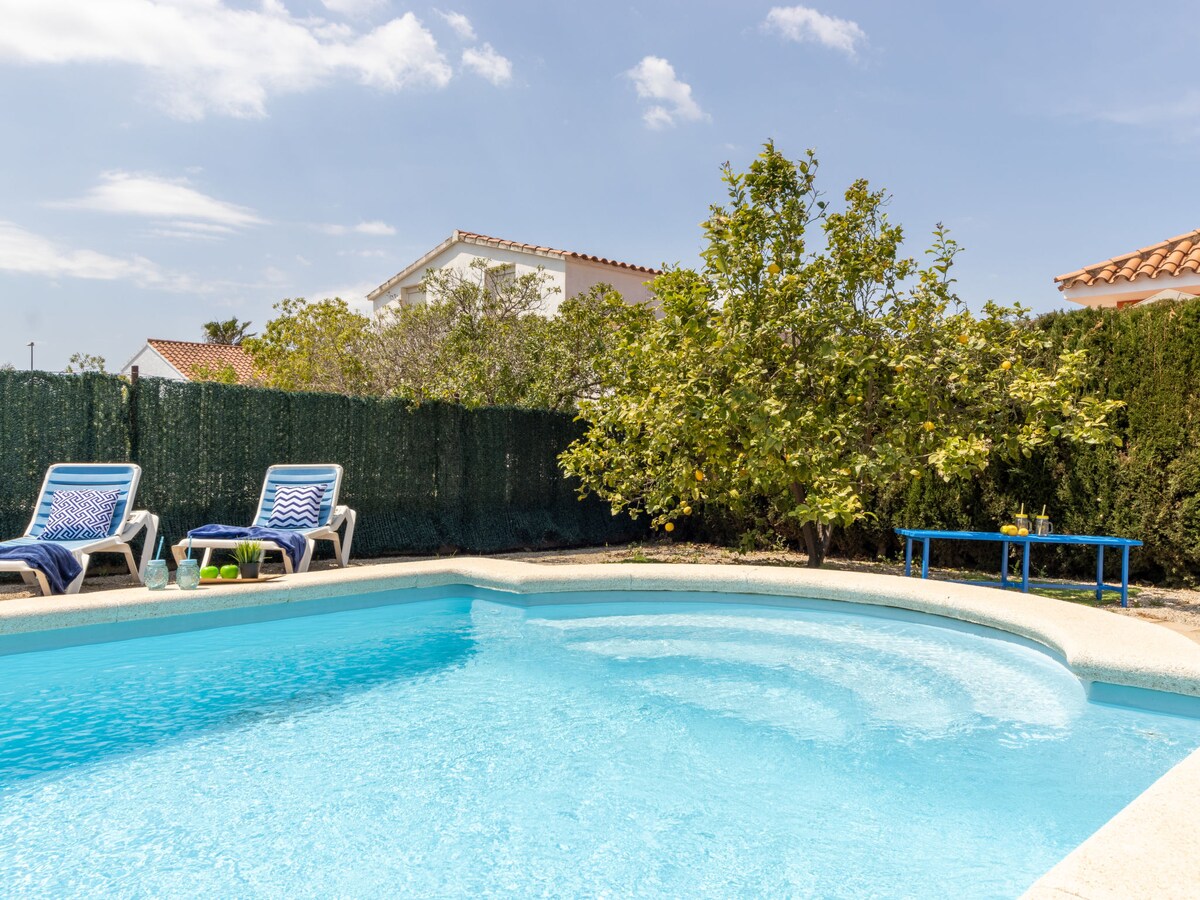 A tranquil swimming pool with a curved shape is surrounded by a well-maintained garden. Two lounge chairs are positioned nearby, alongside a small blue table. Lush greenery and a fruit tree provide a natural backdrop, creating a private outdoor retreat.