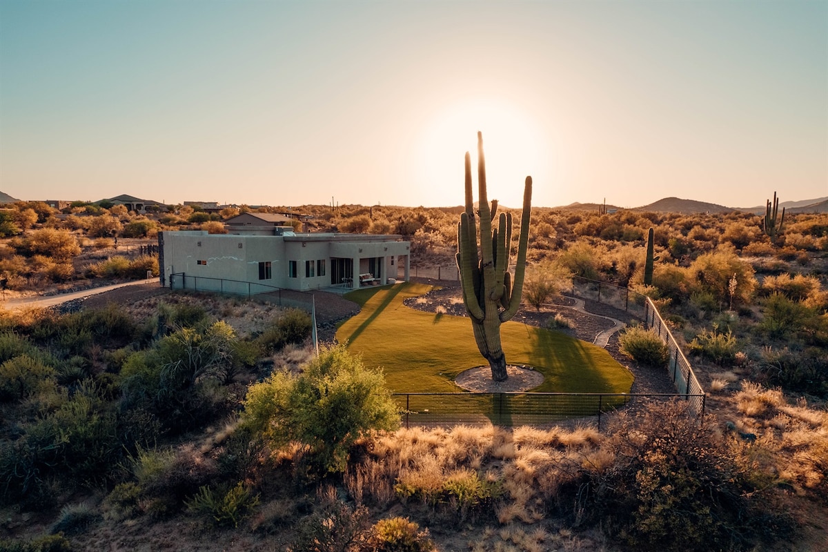 A spacious villa is set against a backdrop of a golden sunset, with a large Saguaro cactus prominently located in the green backyard. The surrounding landscape features various desert flora, creating a serene atmosphere.