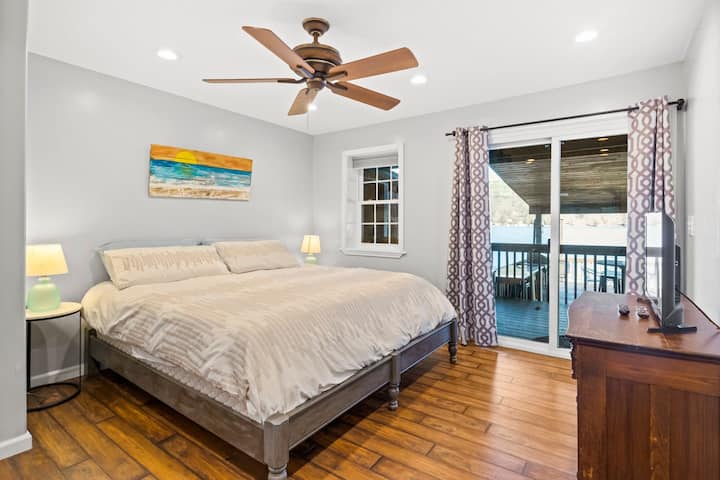 Безупречна спалня с девствено спално бельо, балкон, предлагащ глътка свеж въздух, успокояващ вентилатор на тавана, функционално нощно шкафче с лампа и стилен скрин за организирано съхранение.