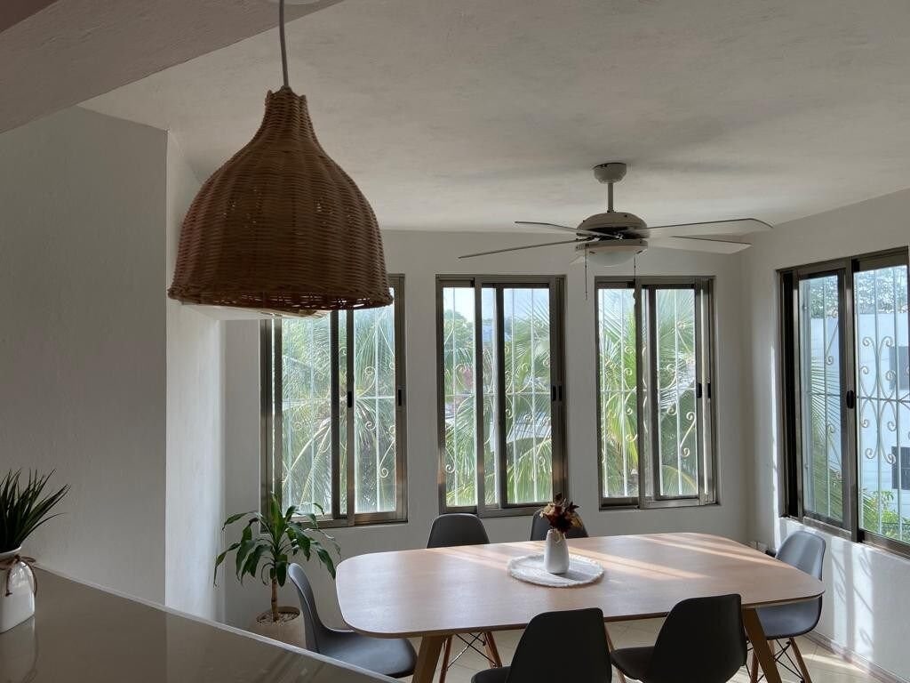 A dining area features a round wooden table surrounded by several chairs, illuminated by natural light from large windows. A ceiling fan provides ventilation, while a decorative pendant lamp hangs above. Green foliage is visible outside, adding a touch of nature to the interior.
