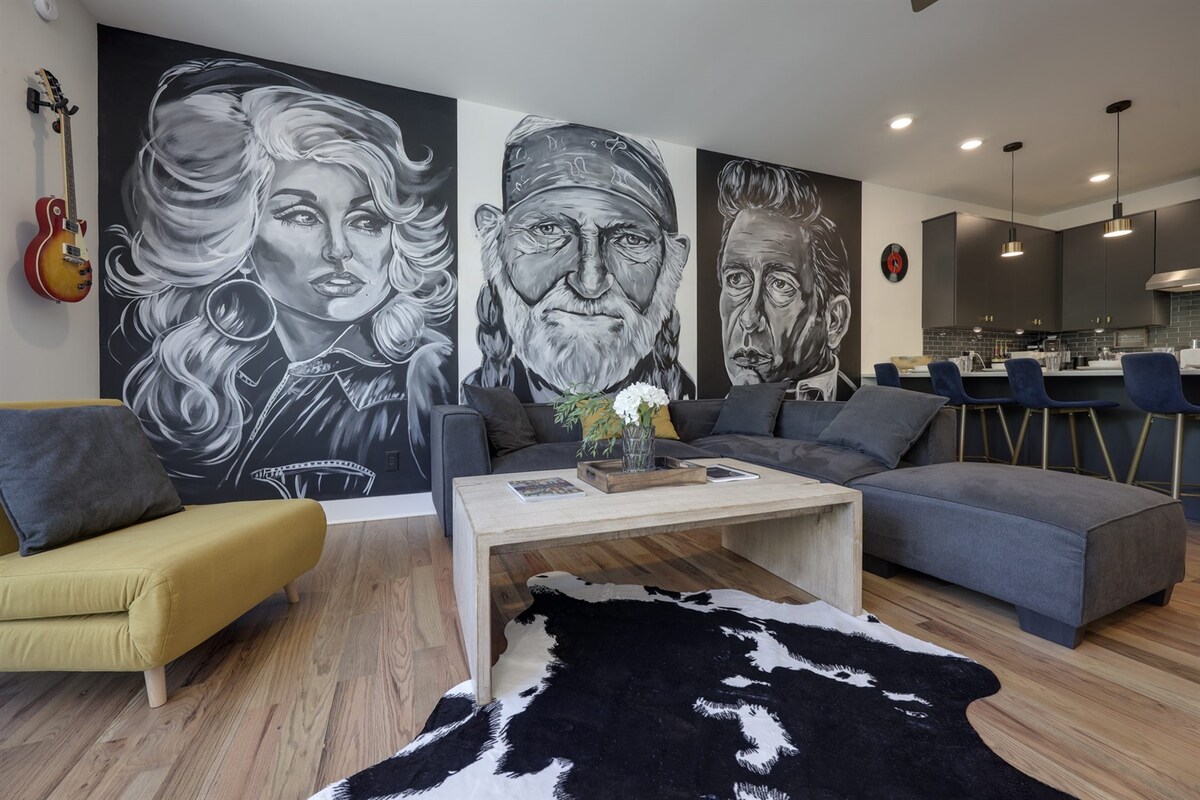 A stylish living area features vibrant wall murals of iconic figures. A large sectional sofa invites relaxation, accompanied by a yellow accent chair. A wooden coffee table sits atop a patterned rug, with a floral arrangement adding a touch of nature to the space. The open kitchen area is visible in the background.