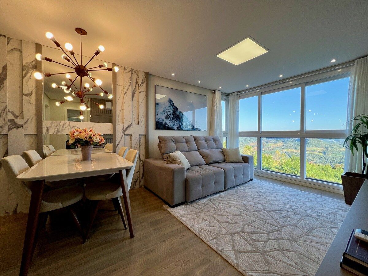 A welcoming living area features a spacious sectional sofa positioned near large windows, offering expansive views of the valley. A dining table for six sits beside the sofa, complemented by a contemporary chandelier overhead. Natural light enhances the soft color palette and elegant décor.