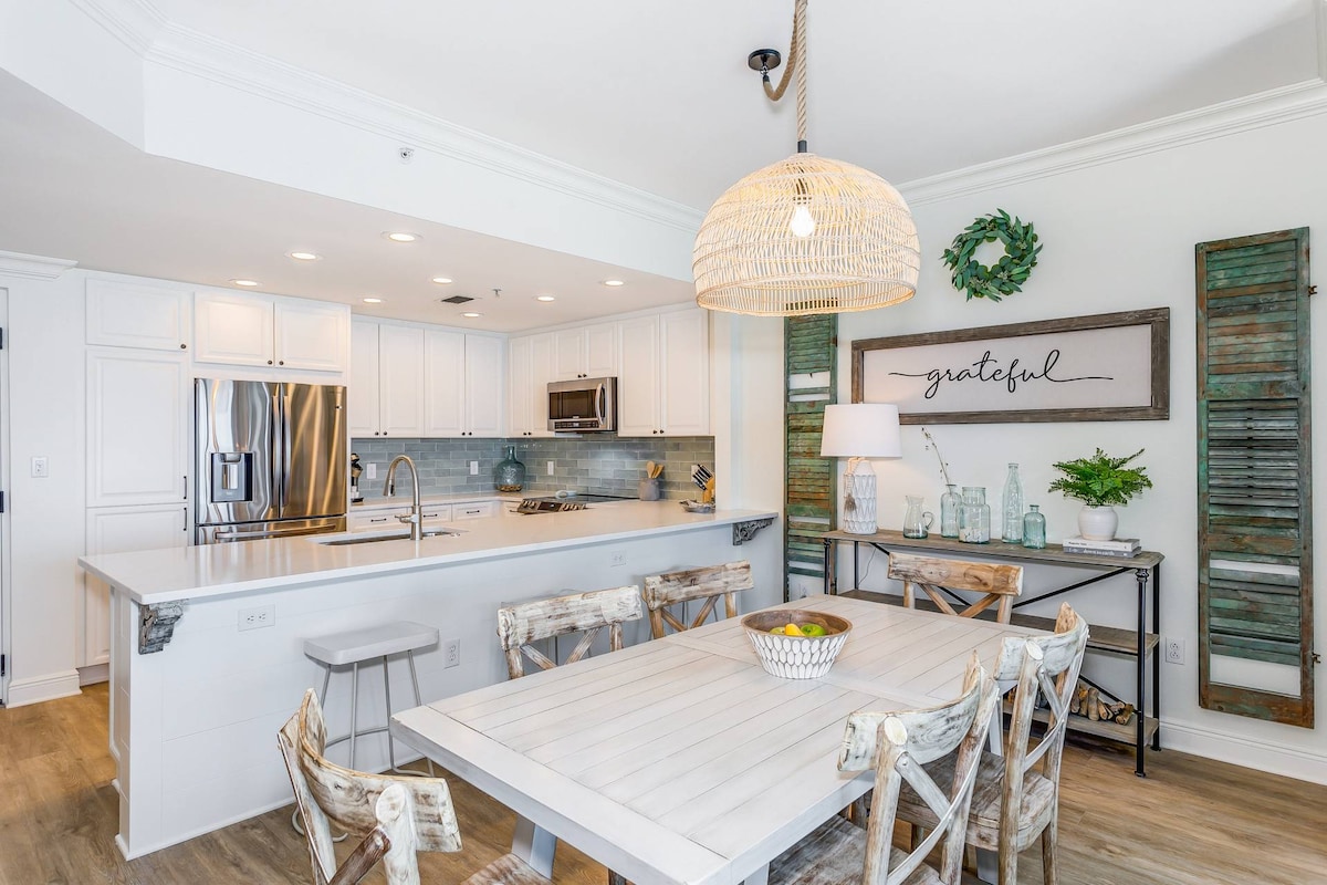 An open kitchen and dining area is displayed, featuring white cabinetry and high-end stainless steel appliances. A large island with a quartz countertop offers additional seating, illuminated by a woven pendant light. A dining table with decorative chairs adds to the coastal aesthetic.