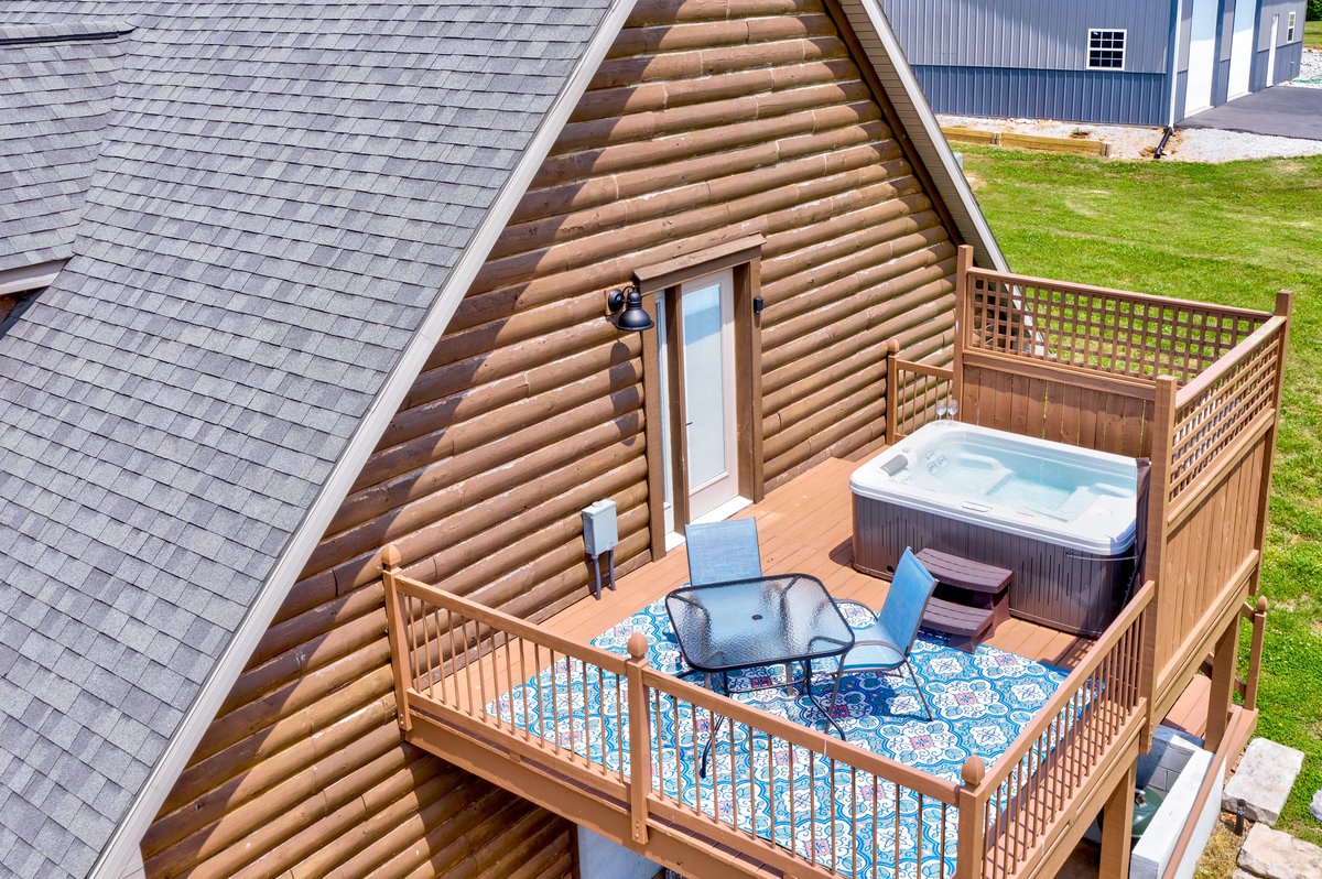 An outdoor deck area features a hot tub, surrounded by wooden railings and a patterned area rug. A small table and two chairs are placed on the deck, offering a relaxing space to enjoy views of the surrounding landscape.