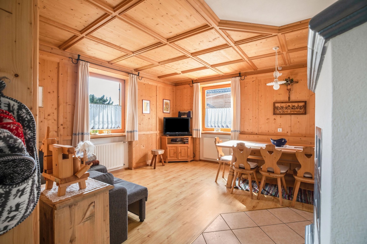 A welcoming living area features a coffered wooden ceiling and natural wood paneling. Two windows allow ample light, illuminating a comfortable seating space and a dining table with multiple chairs. A television is positioned for convenience in the cozy atmosphere.