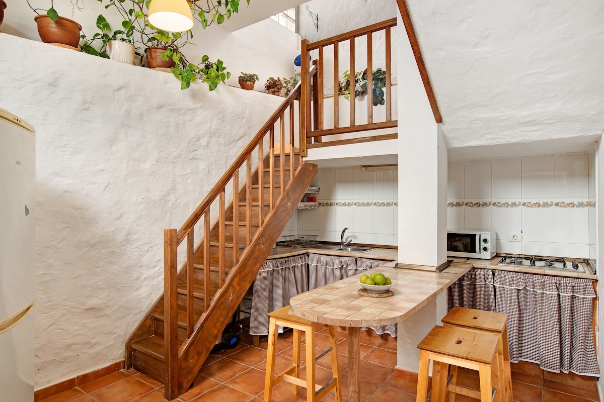 An open kitchen area is presented with a staircase leading to the upper level. A wooden table with stools is positioned, complementing the stone countertops. Potted plants add a touch of greenery, while appliances are arranged neatly within the kitchen space.