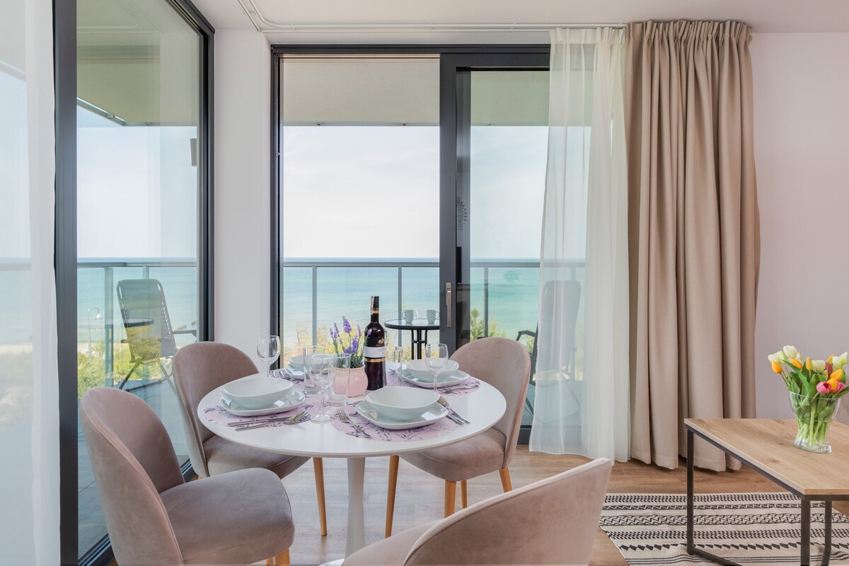 A dining area is presented with a circular table set for four, overlooking a scenic sea view through glass doors. Soft armchairs surround the table, which is elegantly adorned with dishes and a bottle of wine. Natural light enhances the inviting space.