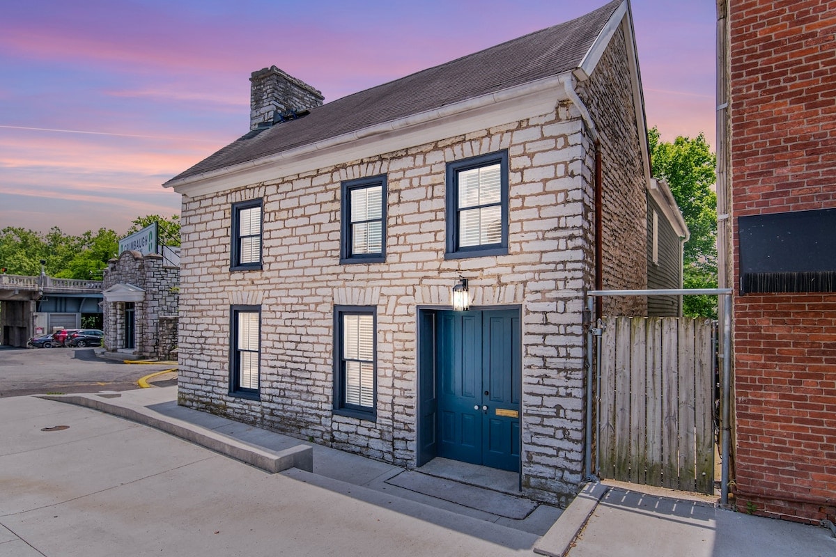 A historic stone house is displayed against a twilight sky, featuring a classic façade with evenly spaced windows and a deep blue front door. Subtle landscaping and a nearby parking area provide a welcoming entrance to this charming property.
