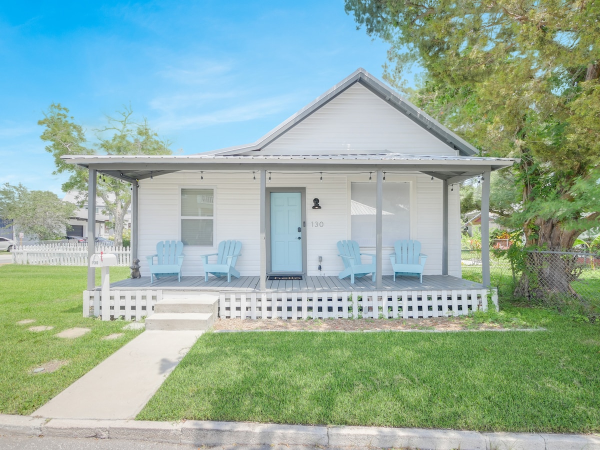 A charming single-story house presents a light-colored exterior, framed by a white picket fence. Two pastel blue adirondack chairs are positioned on the front porch, accompanied by a welcoming front door. Lush green grass complements the inviting entrance.