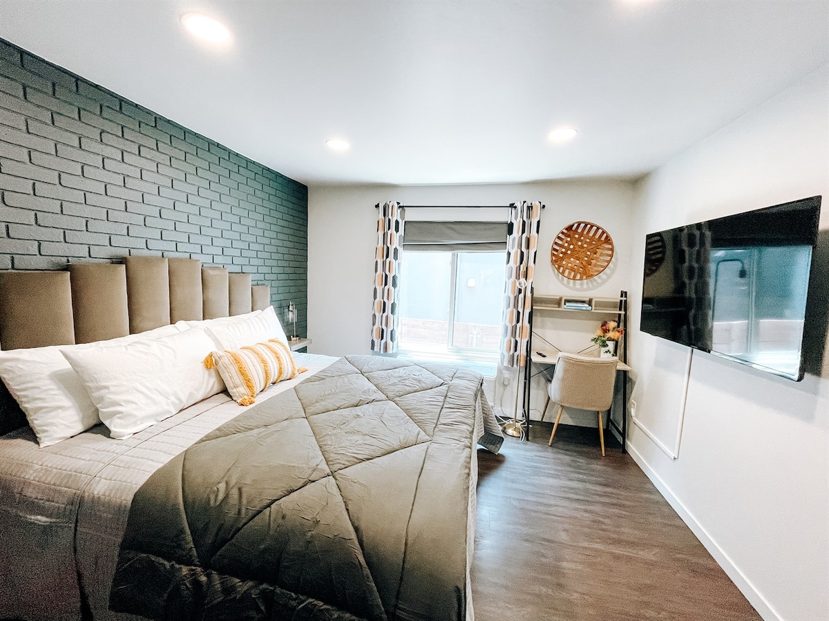 A well-lit bedroom features a spacious king-size bed with a patterned throw blanket. The backdrop consists of textured dark brick walls, while natural light filters through a large window dressed with modern curtains. A small desk with a chair and wall-mounted TV complete the room.