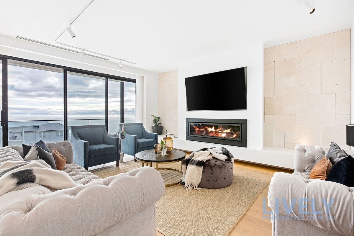 A modern living area features a cozy seating arrangement with plush armchairs and a soft sofa. A large flat-screen TV is mounted above a sleek gas fireplace. Floor-to-ceiling windows provide expansive views, while a neutral-toned rug anchors the furnishings.