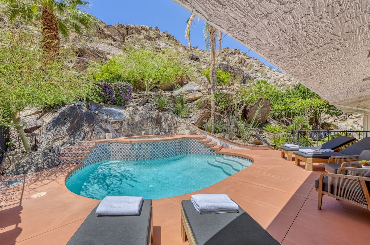 A serene outdoor pool area is framed by natural rock formations and lush tropical landscaping. The pool features a tiled edge and is surrounded by sun loungers with neatly folded towels. The space is designed for relaxation under the clear blue sky.