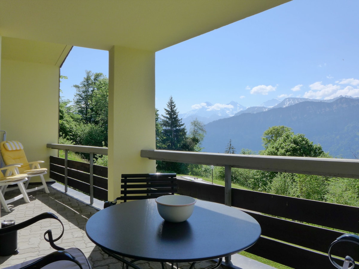 A spacious balcony is visible, featuring outdoor furniture that includes a round table and chairs. The view extends to majestic mountains under a clear blue sky, framed by trees and greenery. Sunlight creates a bright and inviting atmosphere on the balcony.