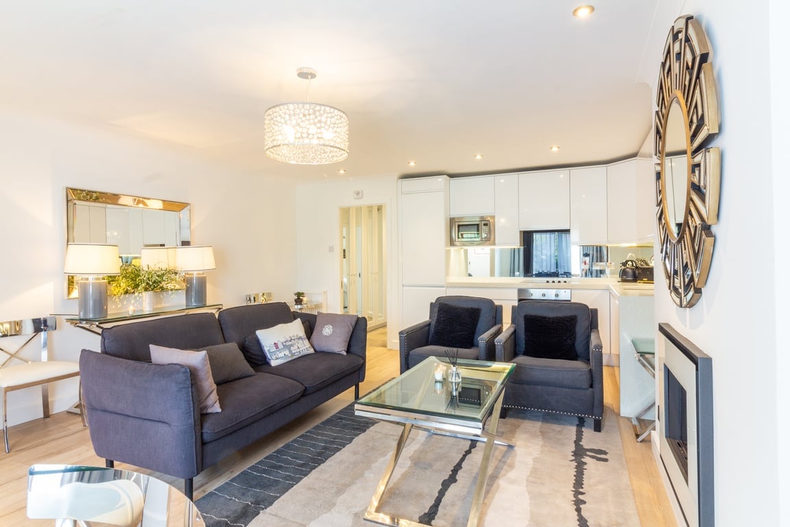 A modern living area is presented with a sleek sofa and two armchairs, complemented by a glass coffee table. Natural light enters through spacious windows, enhancing the contemporary decor, which includes a stylish mirror and an elegant chandelier.
