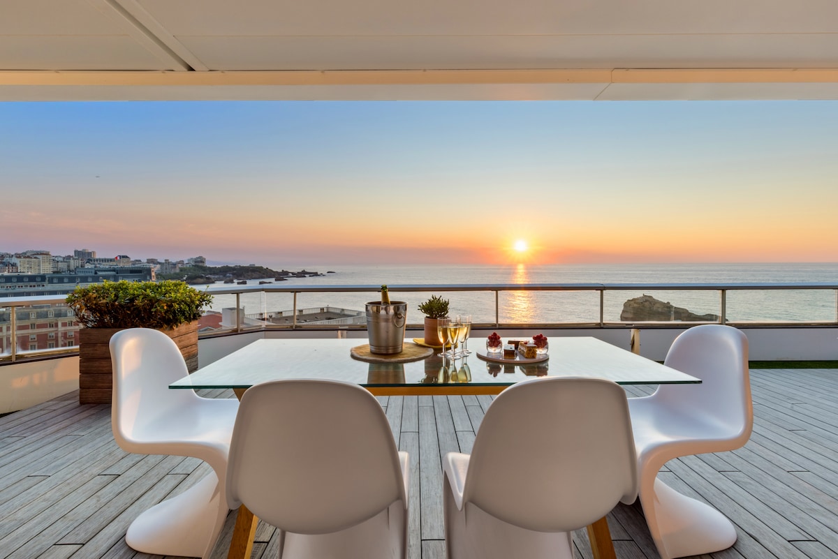 A dining table is set on a spacious terrace, facing a sunset over the ocean. Clear glasses and a bottle of champagne are displayed, surrounded by elegant white chairs. The wooden deck adds warmth to the outdoor setting, enhancing the panoramic view of the seaside.